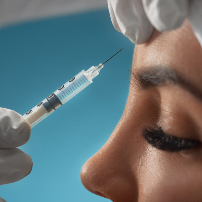 Close up of cosmetic botox injection in female forehead. Side view of cosmetologist using syringe with special liquid and holding patients skin, isolated on blue. Concept of cosmetology, beauty.