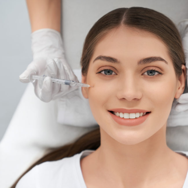 Top view portrait of smiling beautiful young woman in white shirt doing procedure for improvements face skin. Concept of special cosmetics injection for anti-aging in beauty salon.